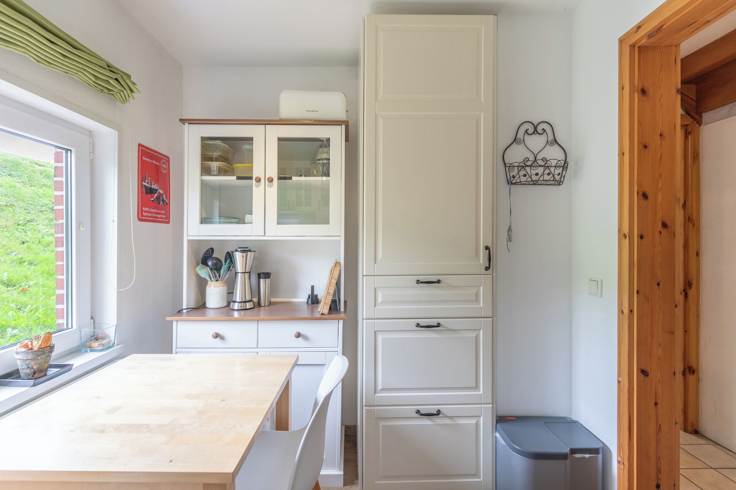 A kitchen with white cabinets, a wooden table, and a window with a view of greenery.
