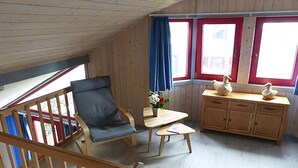 A cozy room with a wooden ceiling, a single window with red trim, a blue curtain, a wooden chair, a small table with a vase of flowers, and a wooden cabinet with decorative items. - Ferienhaus im Ferienpark Mirow (Granzow)