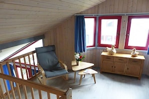 A cozy room with a wooden ceiling, a single window with red trim, a blue curtain, a wooden chair, a small table with a vase of flowers, and a wooden cabinet with decorative items. - Ferienhaus im Ferienpark Mirow (Granzow)