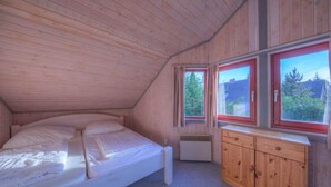 Quarto com teto de madeira, uma cama, um pequeno armário e janelas com vista para árvores e um edifício.