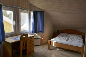 Ett rum med ett skrivbord, en stol och en säng, alla tillverkade av trä. Det finns två fönster med gardiner, och en liten värmare syns.