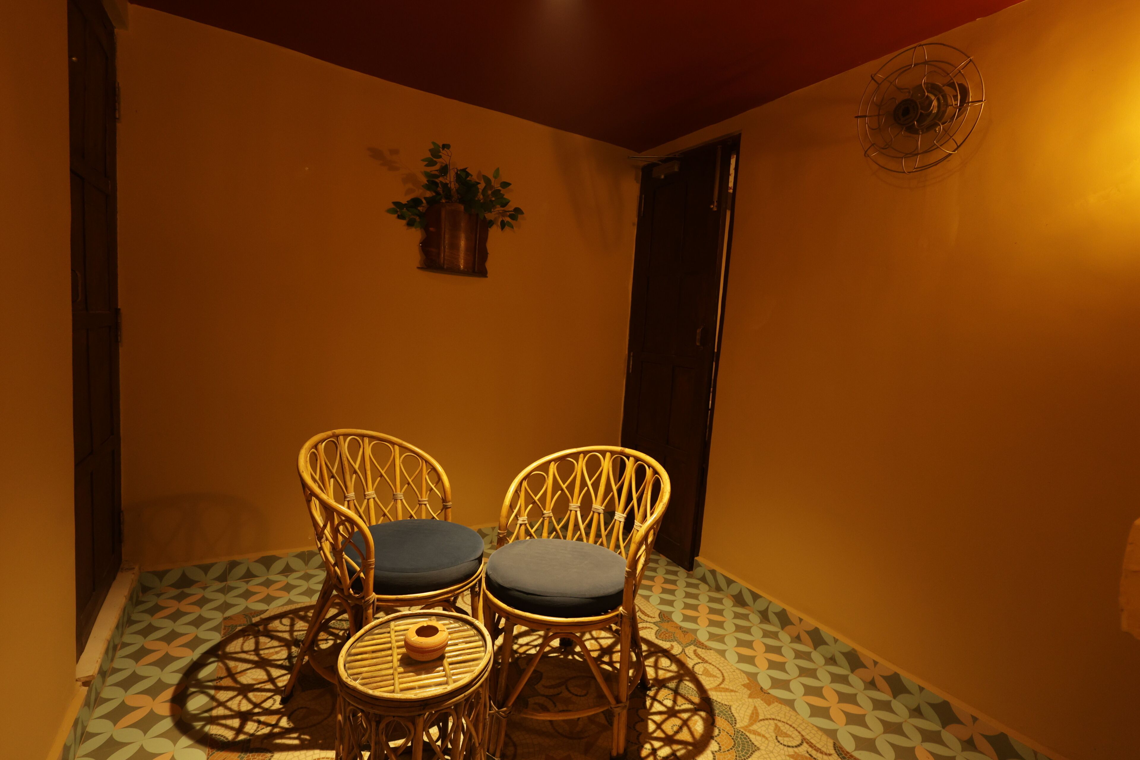 A small room with two wicker chairs and a small table, a potted plant on the wall, and a patterned floor.