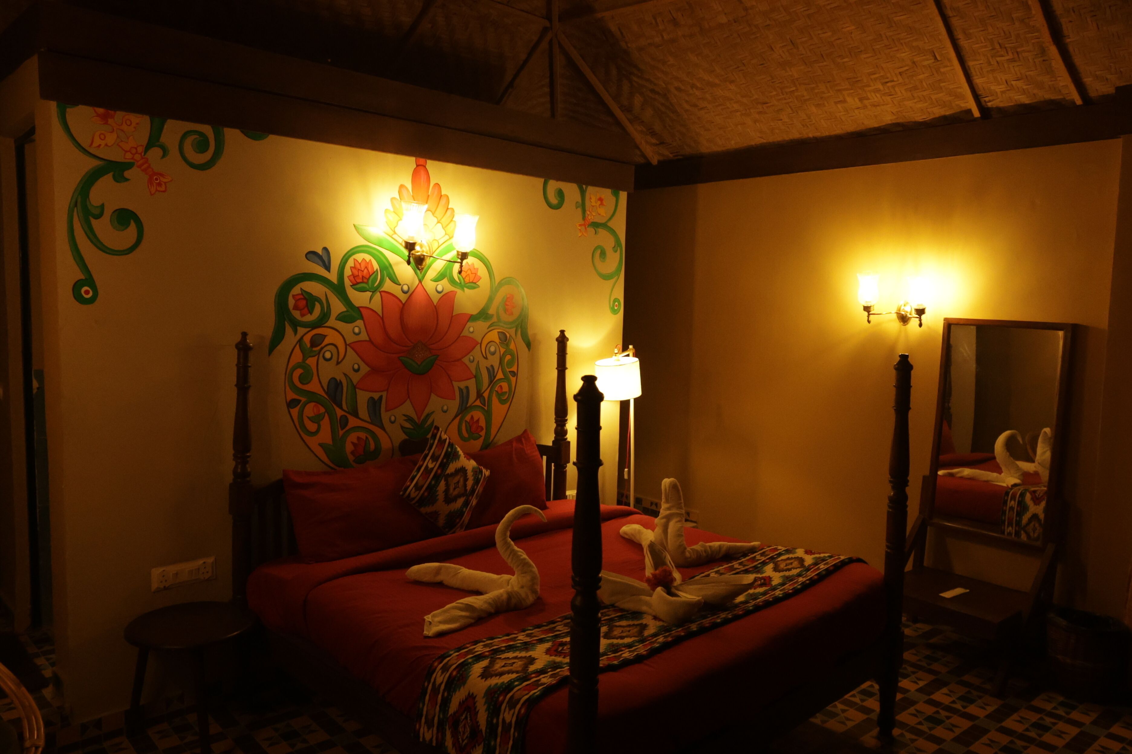 Une chambre avec un lit, une table de chevet, une lampe, un miroir et un mur orné d’une fresque florale.