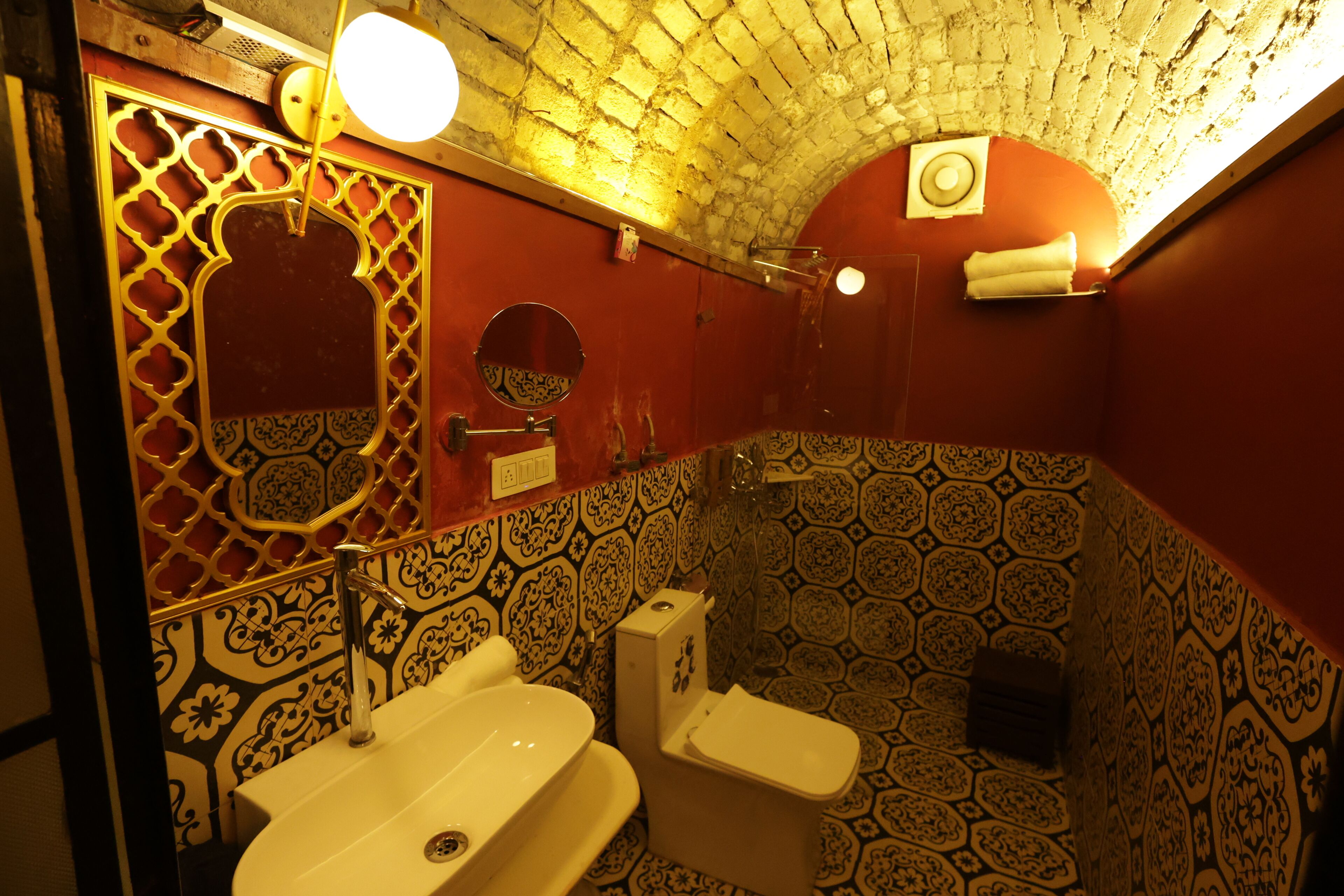 Baño con lavamanos, inodoro y espejo. Las paredes tienen azulejos con diseño, y hay una celosía decorativa sobre el espejo.