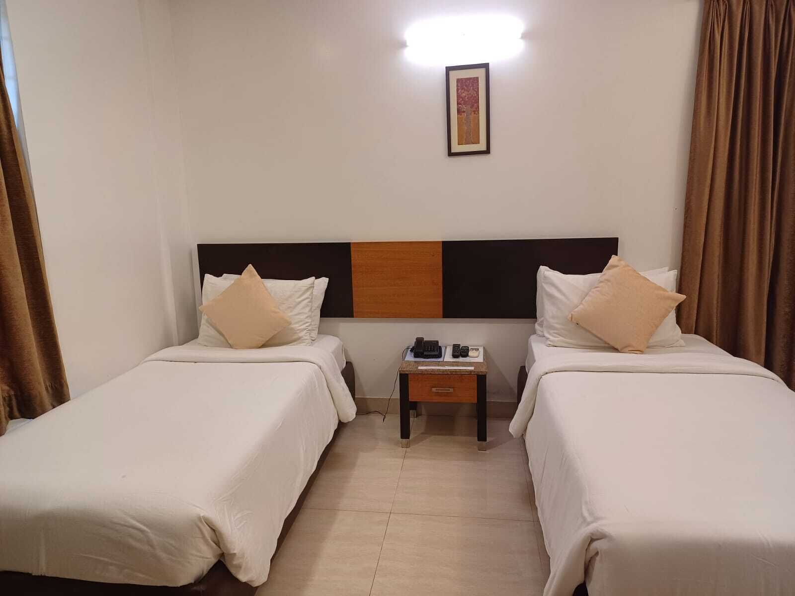 A hotel room with two single beds, a small bedside table with a phone and remote, and a framed picture on the wall.