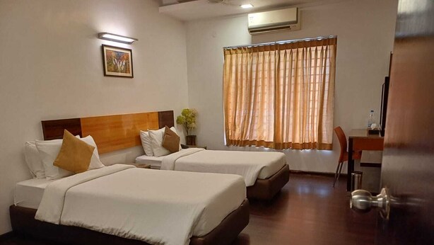 Habitación de hotel con dos camas, un escritorio, una silla, un equipo de aire acondicionado y una ventana con cortinas.