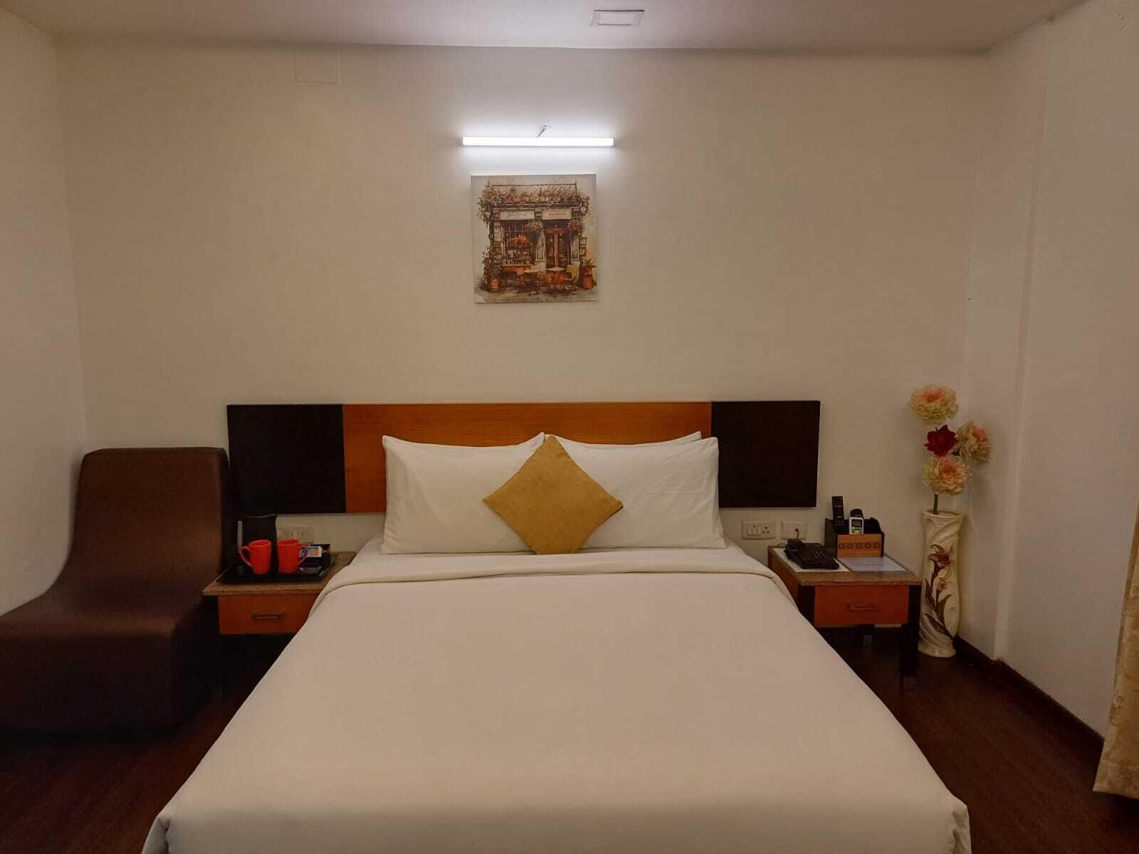 A hotel room with a large bed, a wooden headboard, a nightstand with a phone and a bottle, a brown armchair, and a wall-mounted artwork.