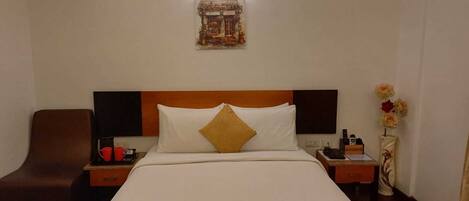 A hotel room with a large bed, a wooden headboard, a nightstand with a phone and a bottle, a brown armchair, and a wall-mounted artwork.