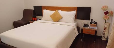 Habitación de hotel con una cama grande, cabecera de madera, mesita de noche con teléfono y lámpara, una silla y una obra de arte en la pared.
