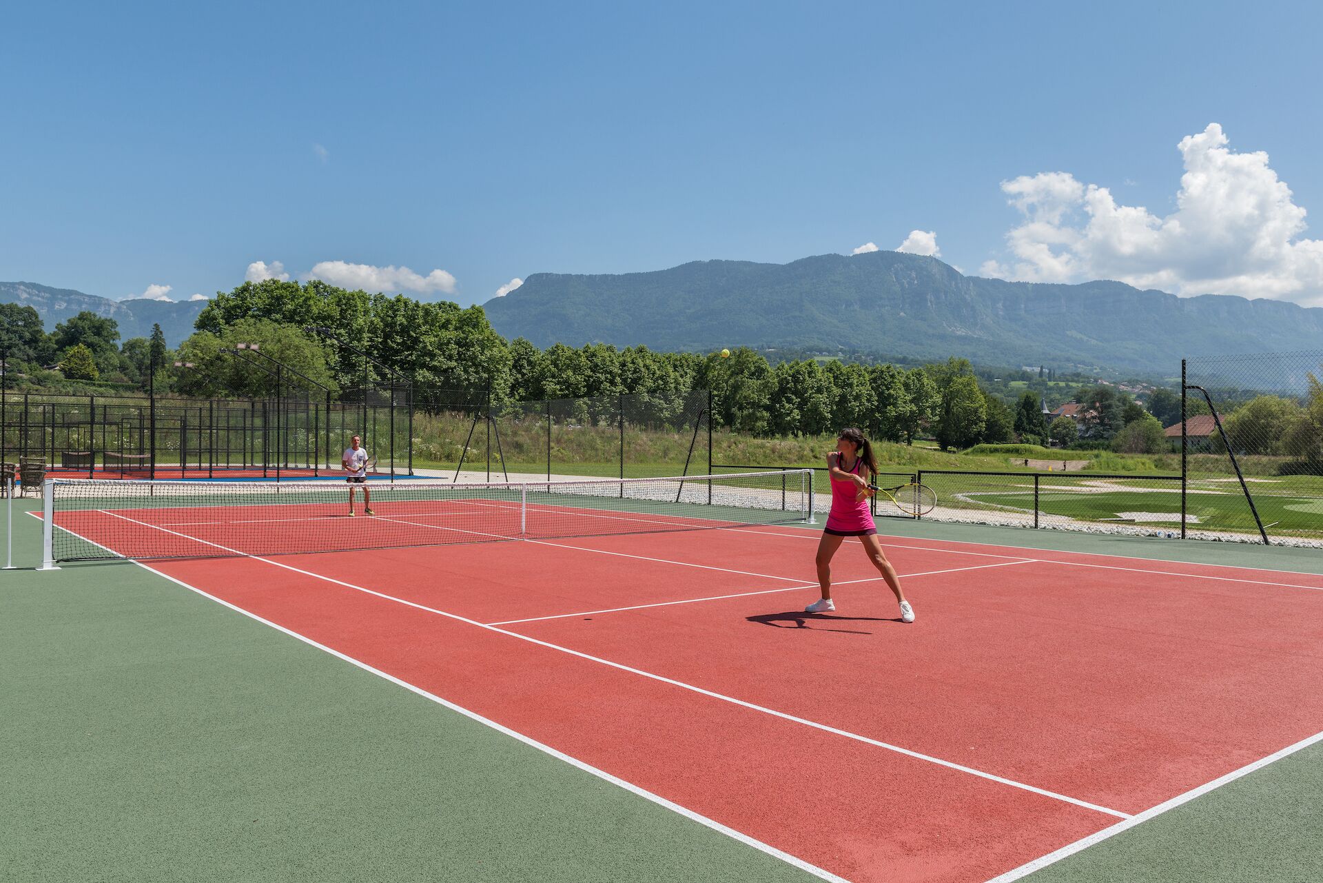 An outdoor tennis court with a net, surrounded by a fence, and mountains in the background.