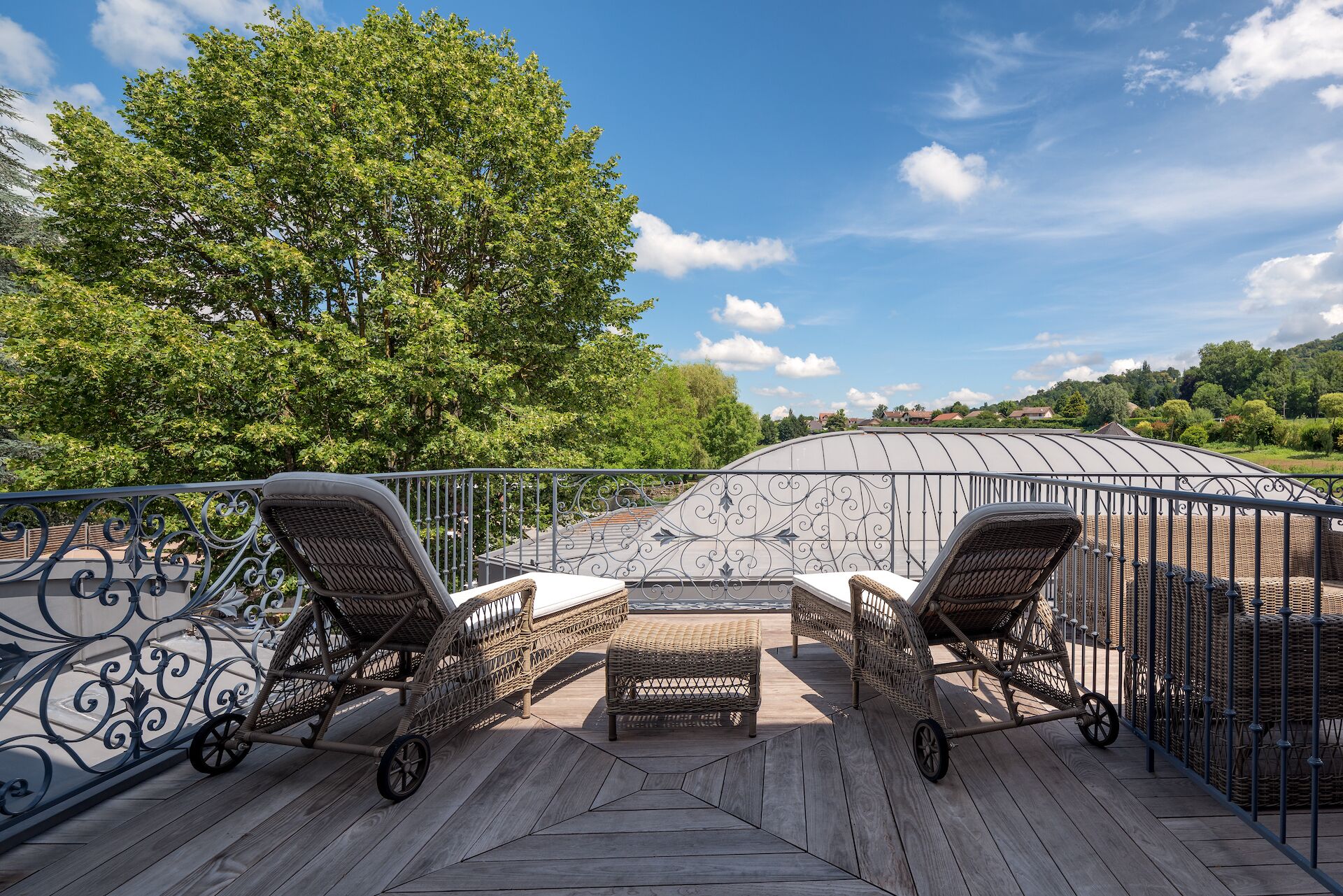 Eine Terrasse mit Korbstühlen und einem kleinen Tisch, umgeben von einem Metallgeländer und einer Glaskonstruktion wie einem Gewächshaus.