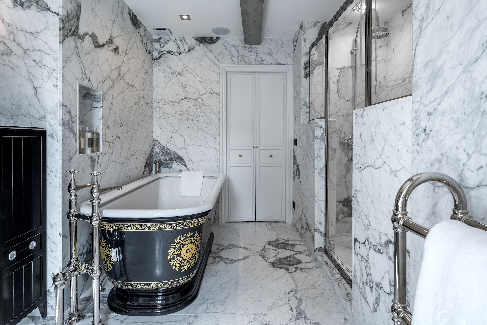 Un baño lujoso con piso de mármol, una bañera independiente con detalles florales dorados, una ducha con mampara de vidrio y una puerta doble blanca.