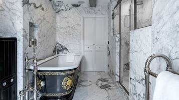Un baño lujoso con piso de mármol, una bañera independiente con detalles florales dorados, una ducha con mampara de vidrio y una puerta doble blanca.