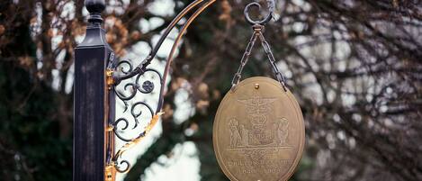 Una placa de latón con texto grabado y el símbolo del caduceo, unida a una estructura de metal negro con ornamentales volutas.