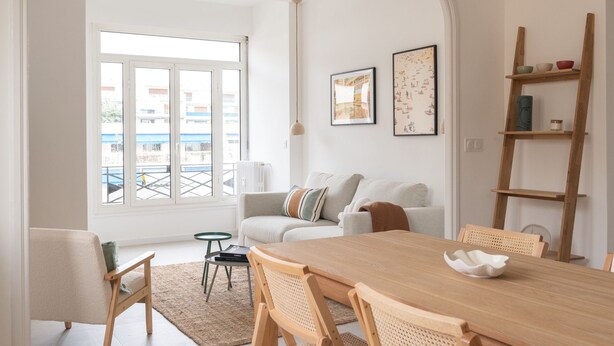 Een ruime woonkamer met een grote houten eettafel, een bank en een salontafel. Er is een raam met uitzicht op gebouwen.