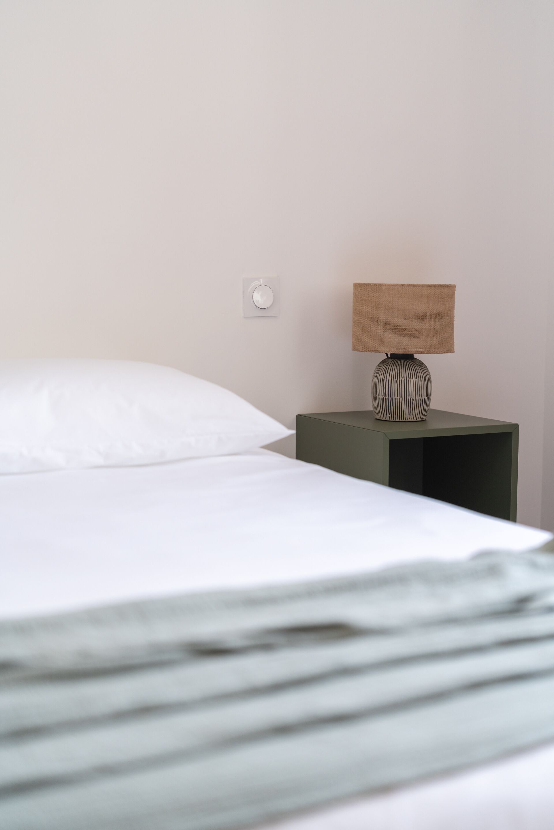 A neatly made bed with white sheets and pillows, a small bedside table with a lamp, and a wall switch.