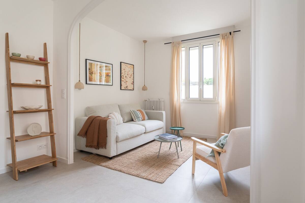 Een woonkamer met een witte bank, een houten ladderrek, een klein rond tafeltje en een stoel. Twee ingelijste foto’s hangen aan de muur, er is een raam met gordijnen en een radiator.