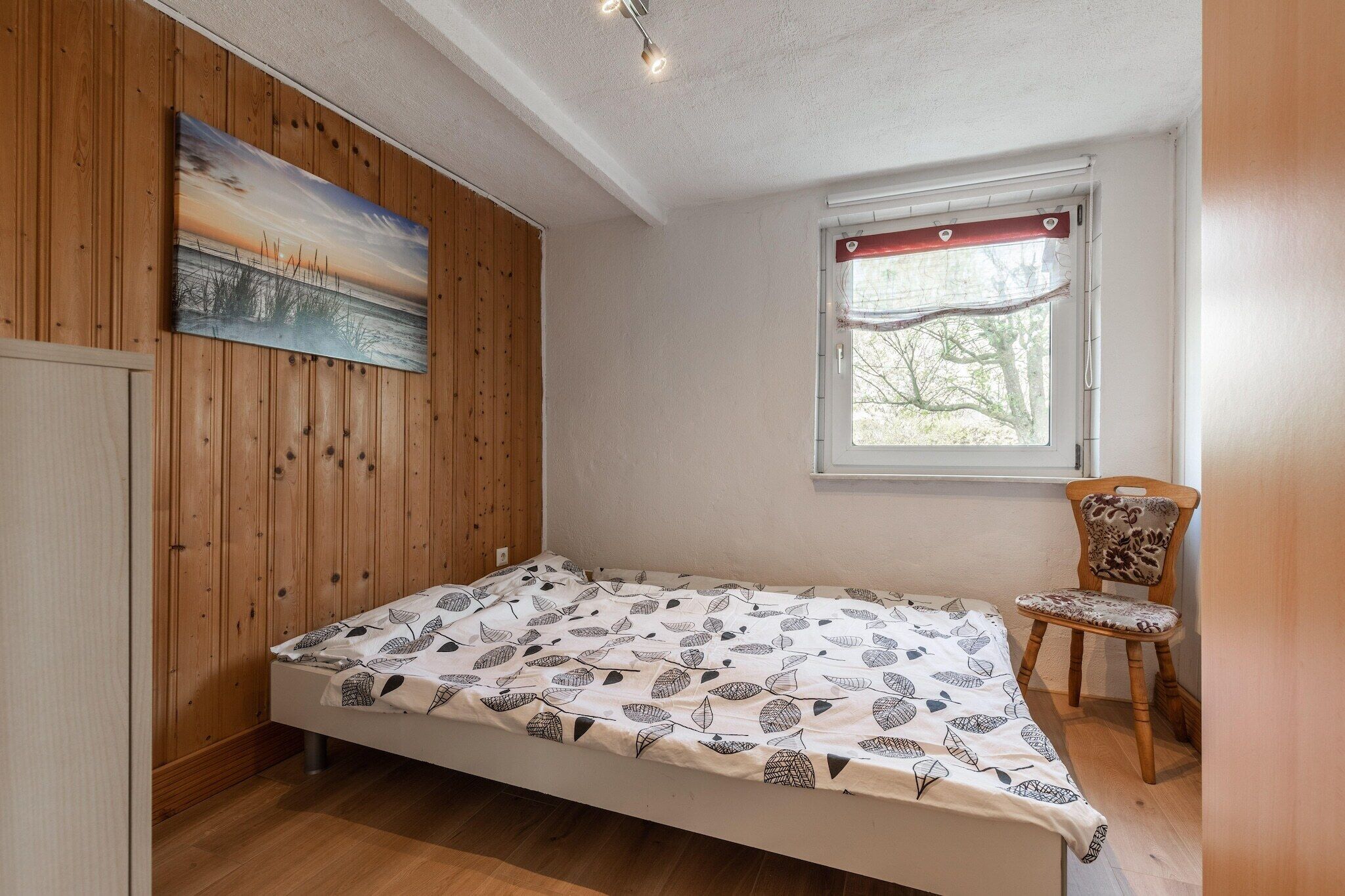 Une petite chambre avec un lit simple, un mur lambrissé, un tableau, une fenêtre donnant sur des arbres et une chaise.