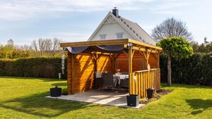 En træterrasse med et bord og stole, omgivet af en hæk og et hus i baggrunden.