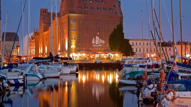 En lystbådehavn med fortøjede både, en stor bygning med et muralmaleri, og et roligt vandareal der spejler omgivelserne.