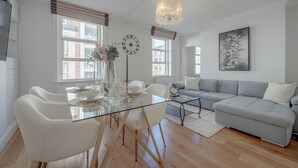 A modern living room with a glass dining table, white chairs, a grey sofa, and a chandelier. - Iconic Oxford Street Apartment ✨ 2 Bed 1 Bath ✨ Tube ✨ Bond Street 📍 (Greater London)
