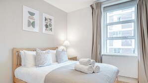 A neatly made bed with a beige quilt, a wooden headboard, and two pillows. There is a nightstand with a lamp and a window with curtains. - Iconic Oxford Street Apartment ✨ 2 Bed 1 Bath ✨ Tube ✨ Bond Street 📍 (Greater London)