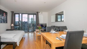 Ein modernes Hotelzimmer mit einem Bett, einem Esstisch für zwei Personen, einer Couch und Blick auf eine Skyline durch große Fenster.