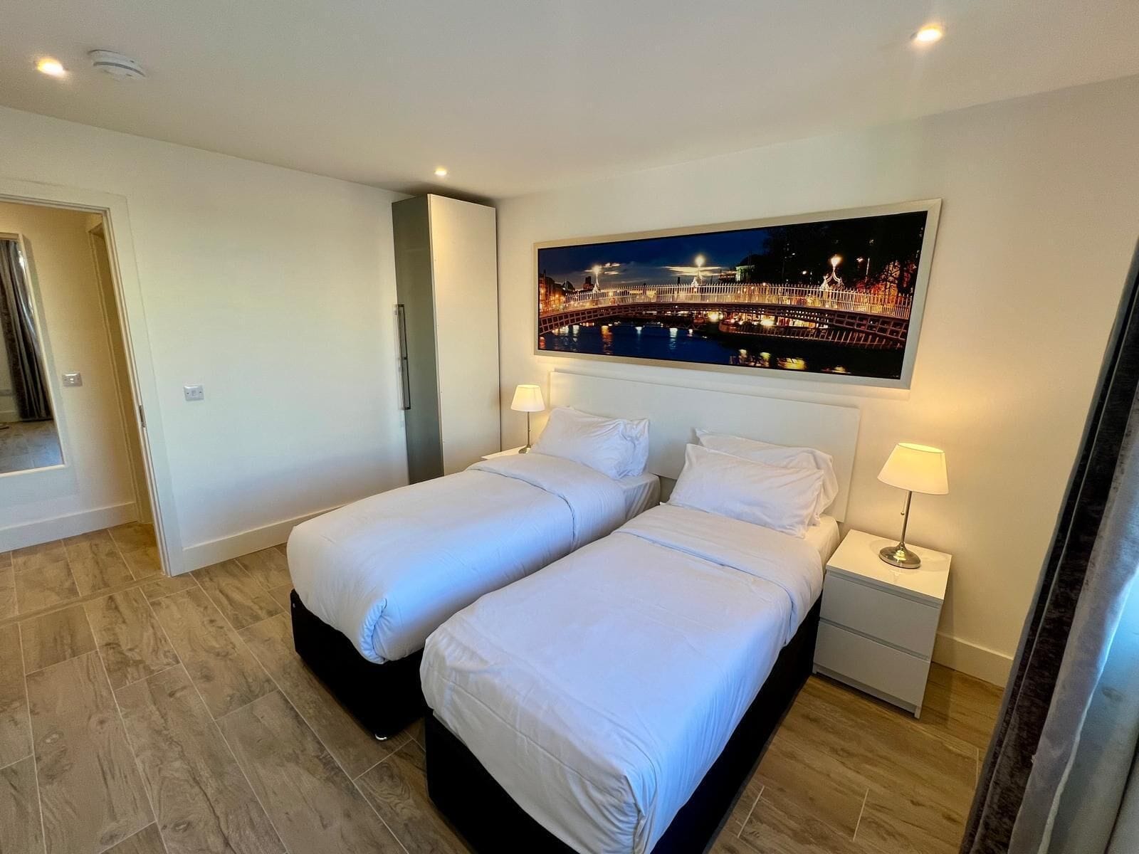 A hotel room with two beds, a nightstand with a lamp, and a large cityscape painting on the wall.