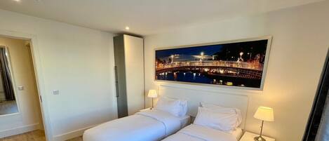 A hotel room with two beds, a nightstand with a lamp, and a large cityscape painting on the wall.