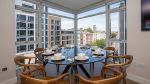 Ein Essbereich mit einem runden Tisch, gedeckt für sechs Personen, umgeben von Holzstühlen, großen Fenstern mit Blick auf die Stadt und einem modernen Gebäude im Hintergrund.