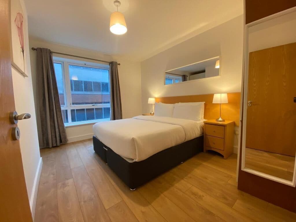 A bedroom with a bed, bedside lamps, a window with curtains, a mirror above the bed, and a wooden door.