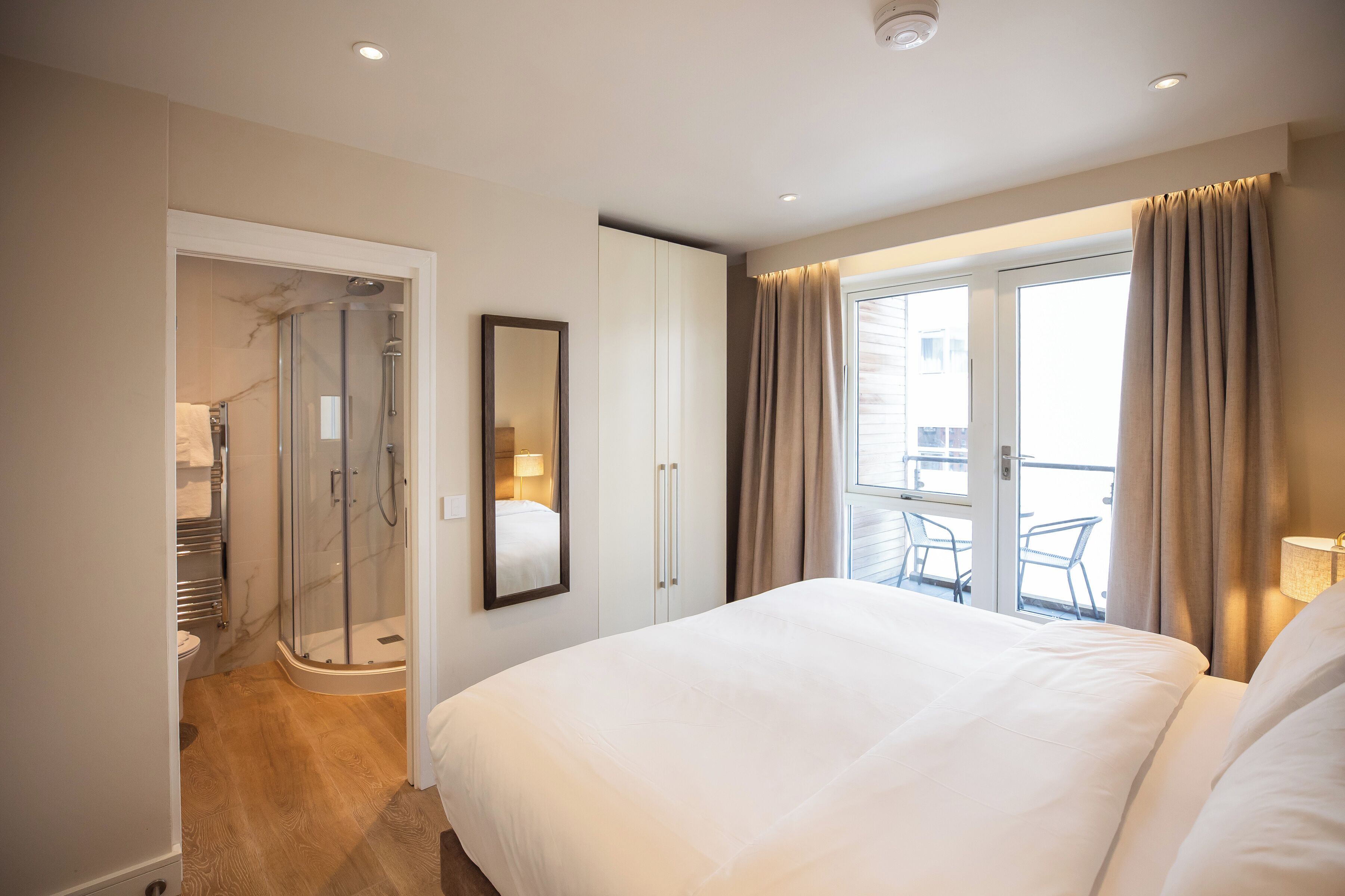 A hotel room with a large bed, a walk-in shower, a balcony with a chair, and a view of the city.