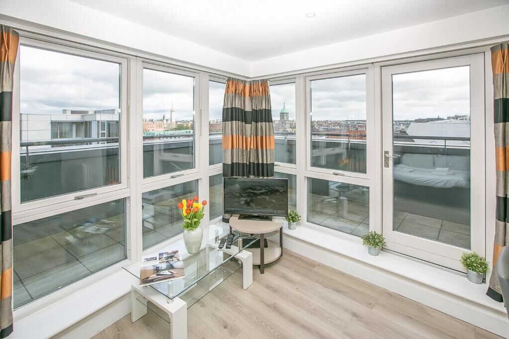 A modern living room with large windows, a flat-screen TV, a glass coffee table, and a small plant.