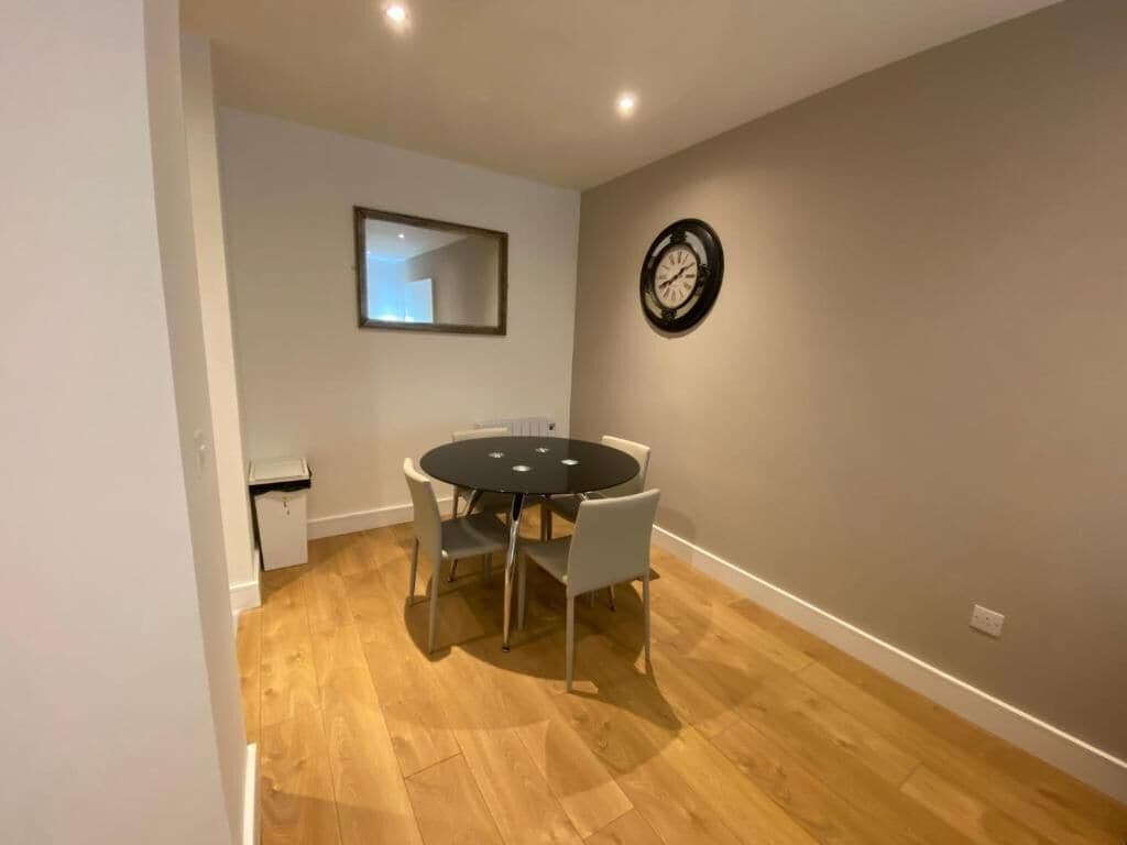 A room with a round dining table, four chairs, a small trash bin, a wall clock, and a mirror.
