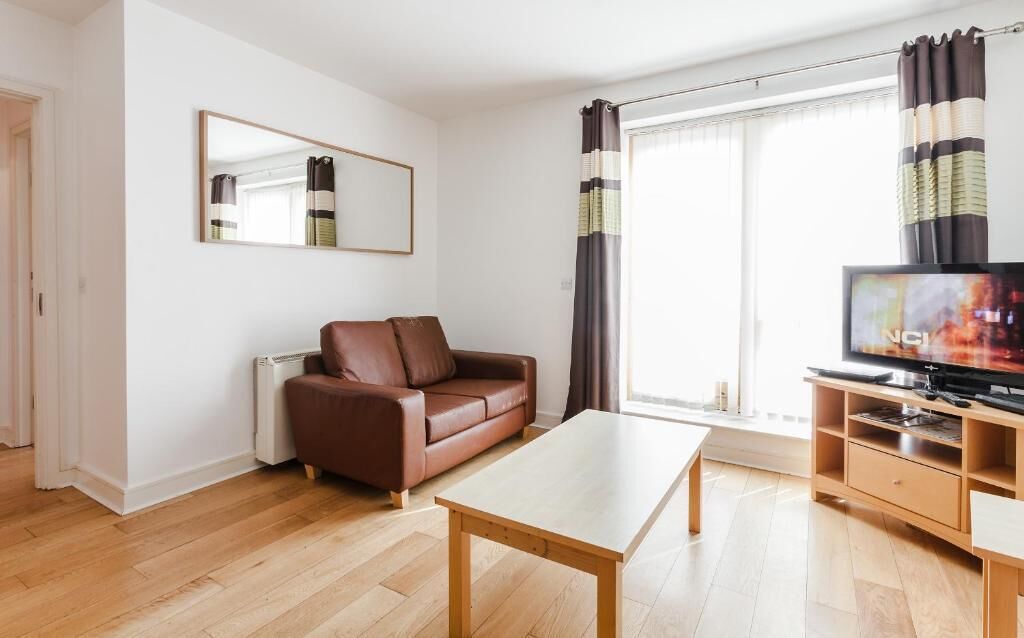 A modern living room with a brown leather sofa, a wooden coffee table, a flat-screen TV on a wooden stand, and a large window with curtains.