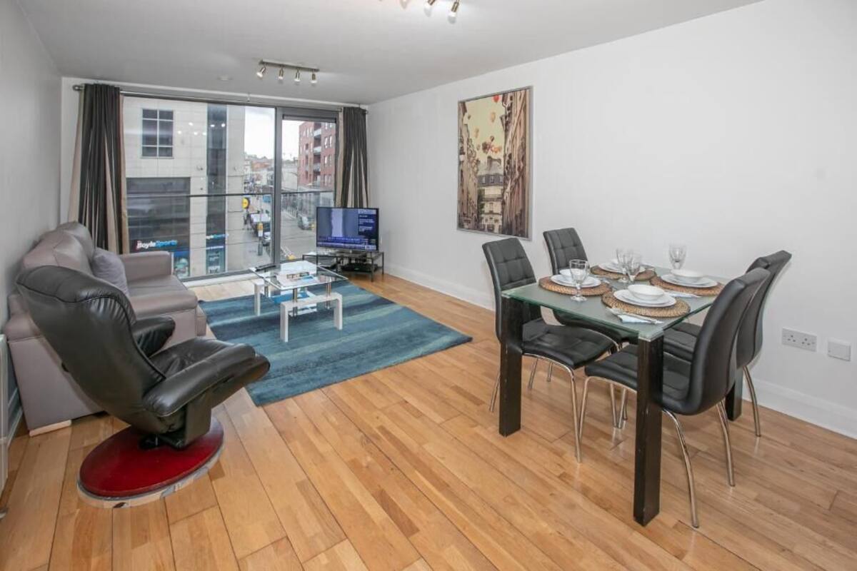 Un salon moderne avec un canapé, une table à manger pour deux personnes, un téléviseur à écran plat et une grande fenêtre donnant sur la ville.