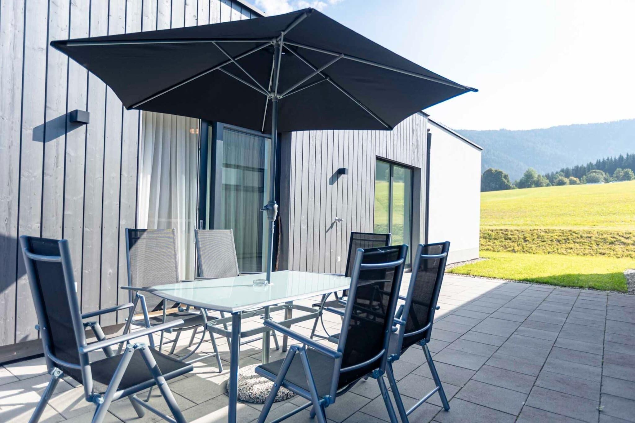 A patio with a glass table and four chairs, an umbrella, and a view of a field and mountains.
