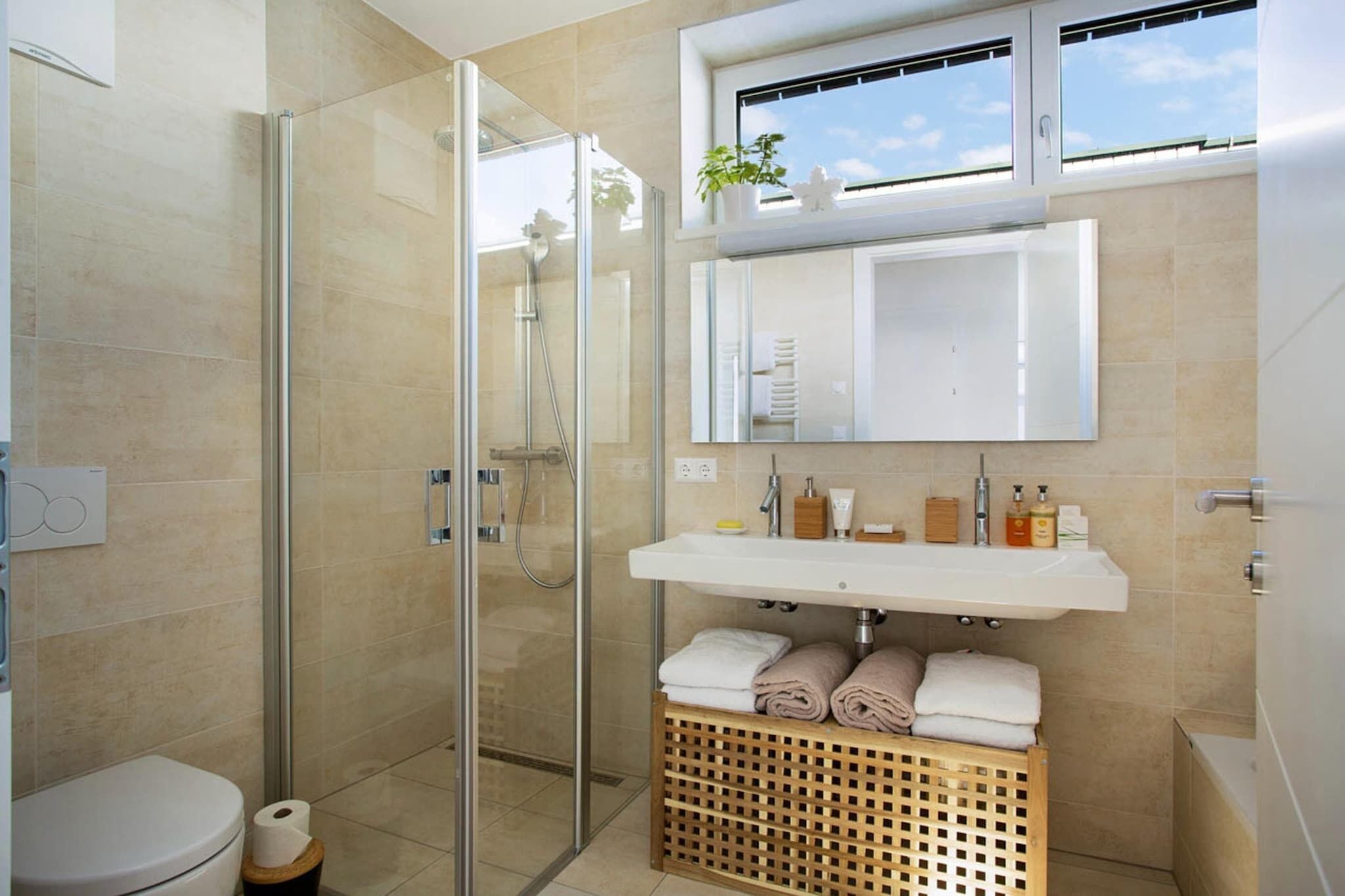 Ein modernes Badezimmer mit einer Duschkabine aus Glas, einem weißen Waschbecken mit Badutensilien und einem Fenster über dem Spiegel.