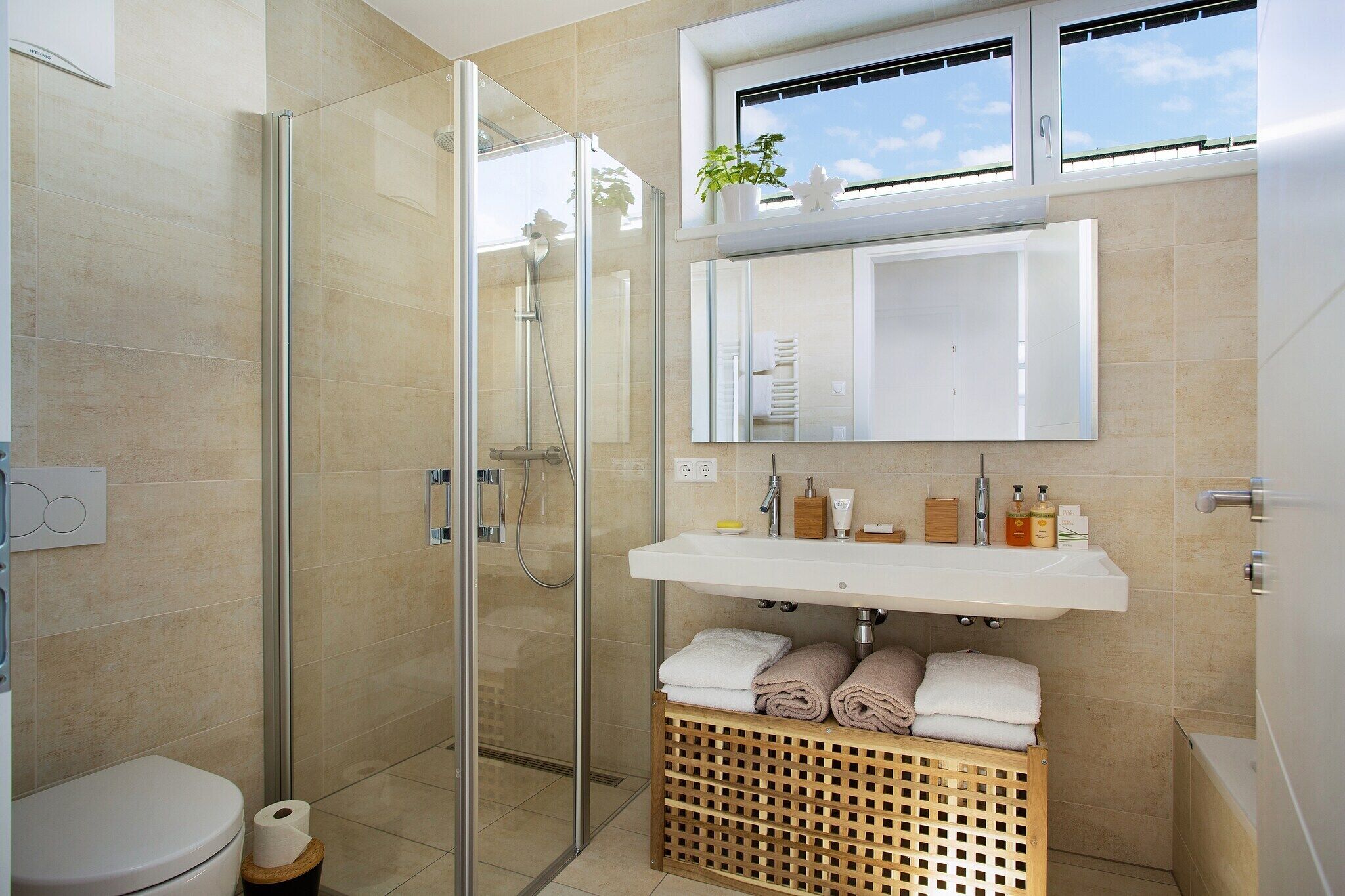 Ein modernes Badezimmer mit einer gläsernen Duschkabine, einem weißen Waschbecken mit Pflegeprodukten und einem Holzkorb mit Handtüchern.