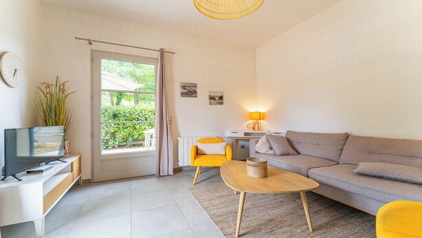 A modern living room with a grey sofa, a yellow armchair, a wooden coffee table, a TV on a white desk, and a window with a view of greenery. - DOMAINE DU FLAMAND - Gite DUNE  (Naujac-sur-Mer)