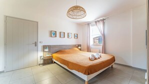 A bedroom with a bed, a nightstand, a lamp, a window with curtains, and a wall with framed pictures. - DOMAINE DU FLAMAND - Gite DUNE  (Naujac-sur-Mer)