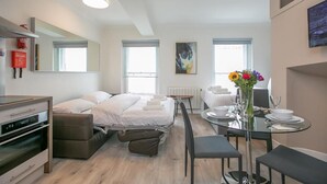 Ein modernes Hotelzimmer mit einem Bett, einem Esstisch mit Stühlen, einer kleinen Küchenzeile und einem großen Fenster.