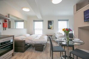 Een moderne hotelkamer met een bed, een eettafel met stoelen, een kleine kitchenette en een groot raam.
