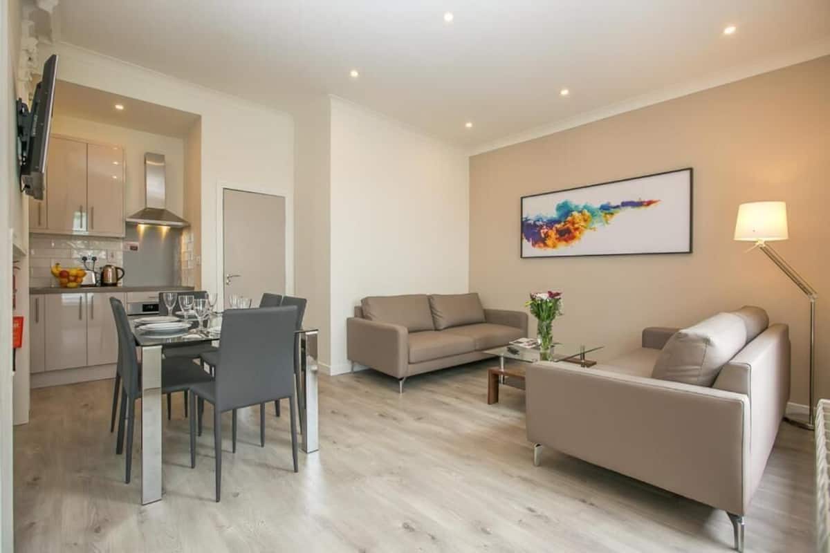 A modern living room with a beige sofa, a dining table set for four, and a kitchen in the background.