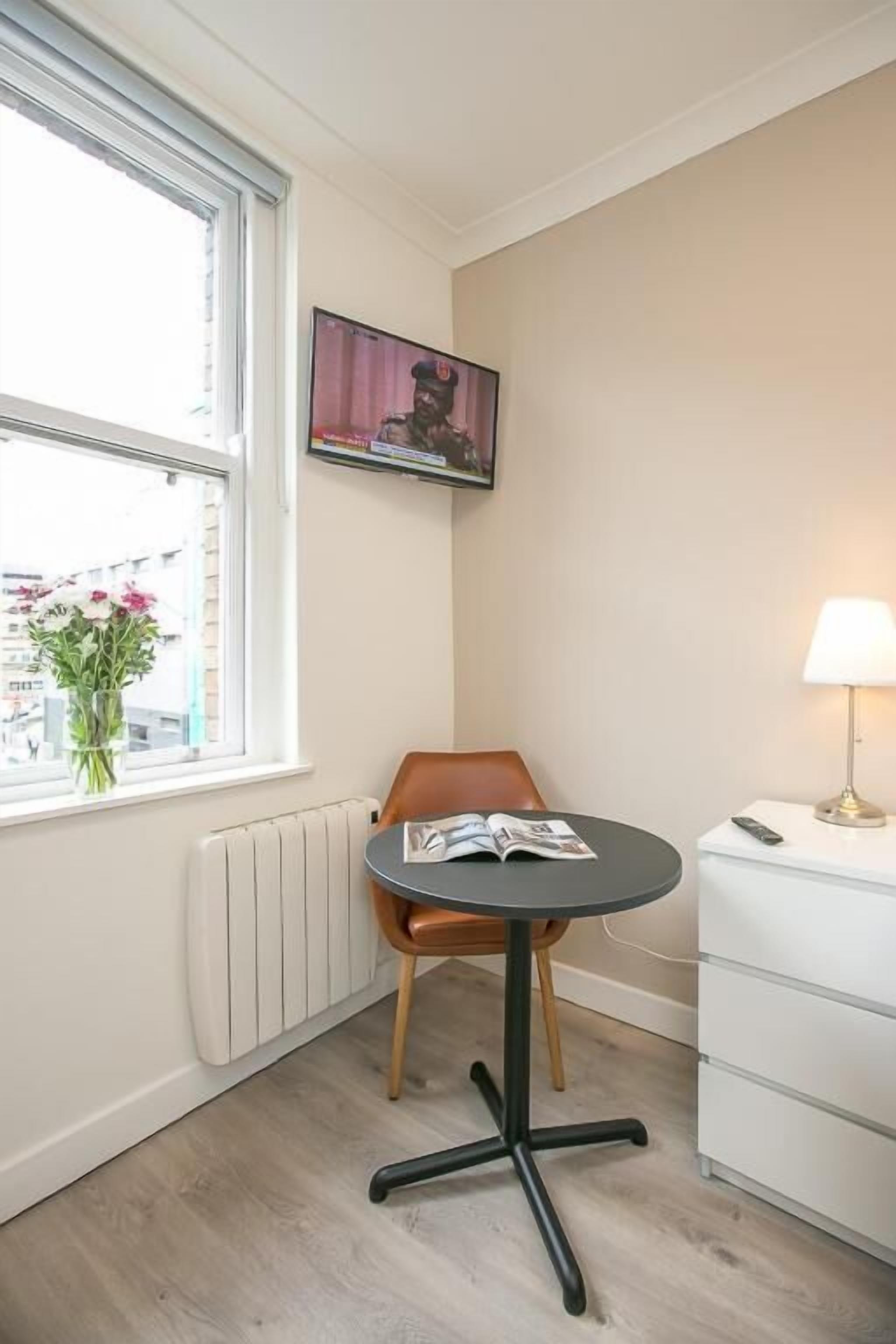 A corner room with a window, a chair, a table, a lamp, and a radiator.