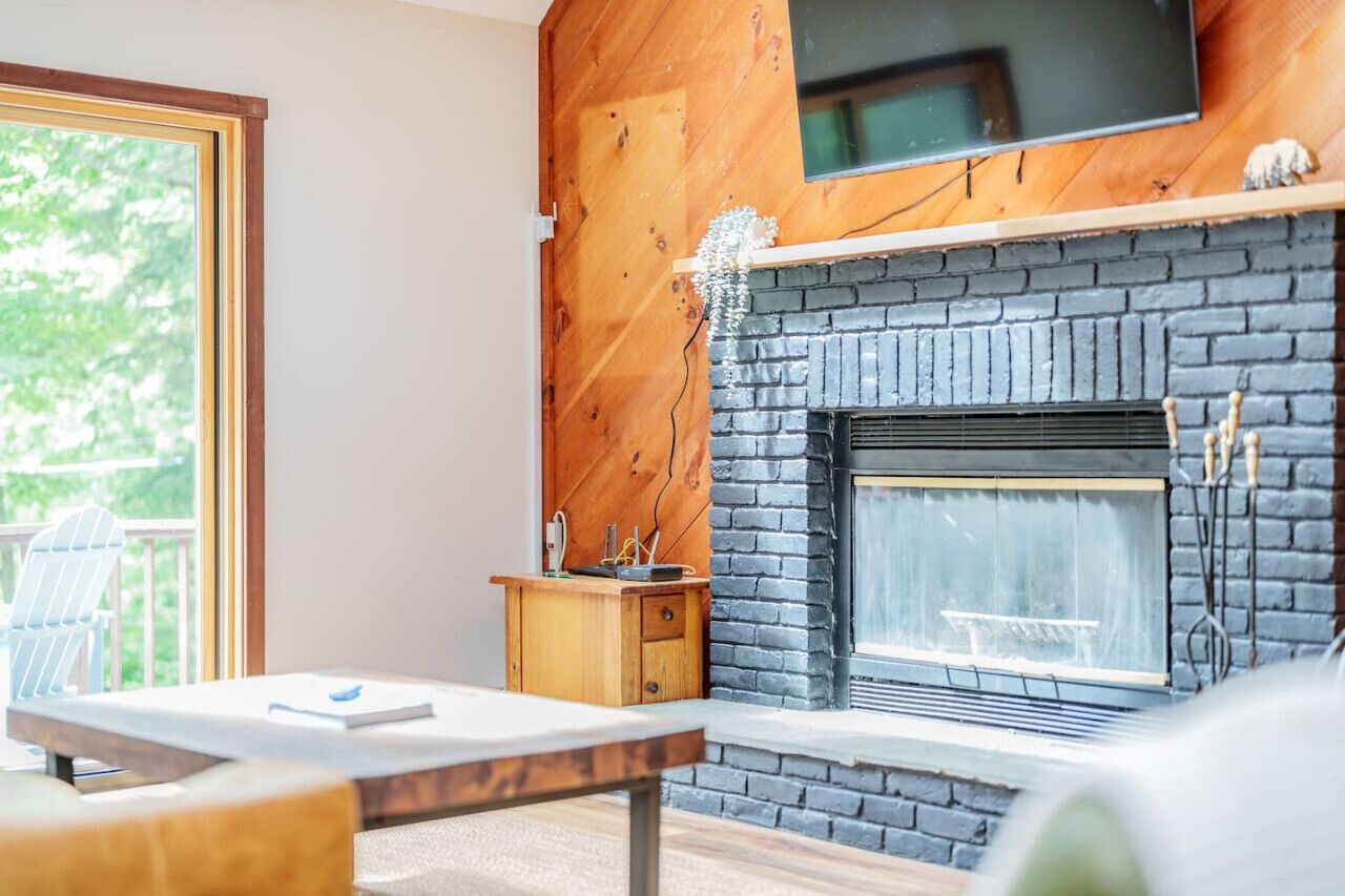 Una sala de estar moderna con chimenea, paneles de madera, un televisor montado encima y vistas a la vegetación exterior.