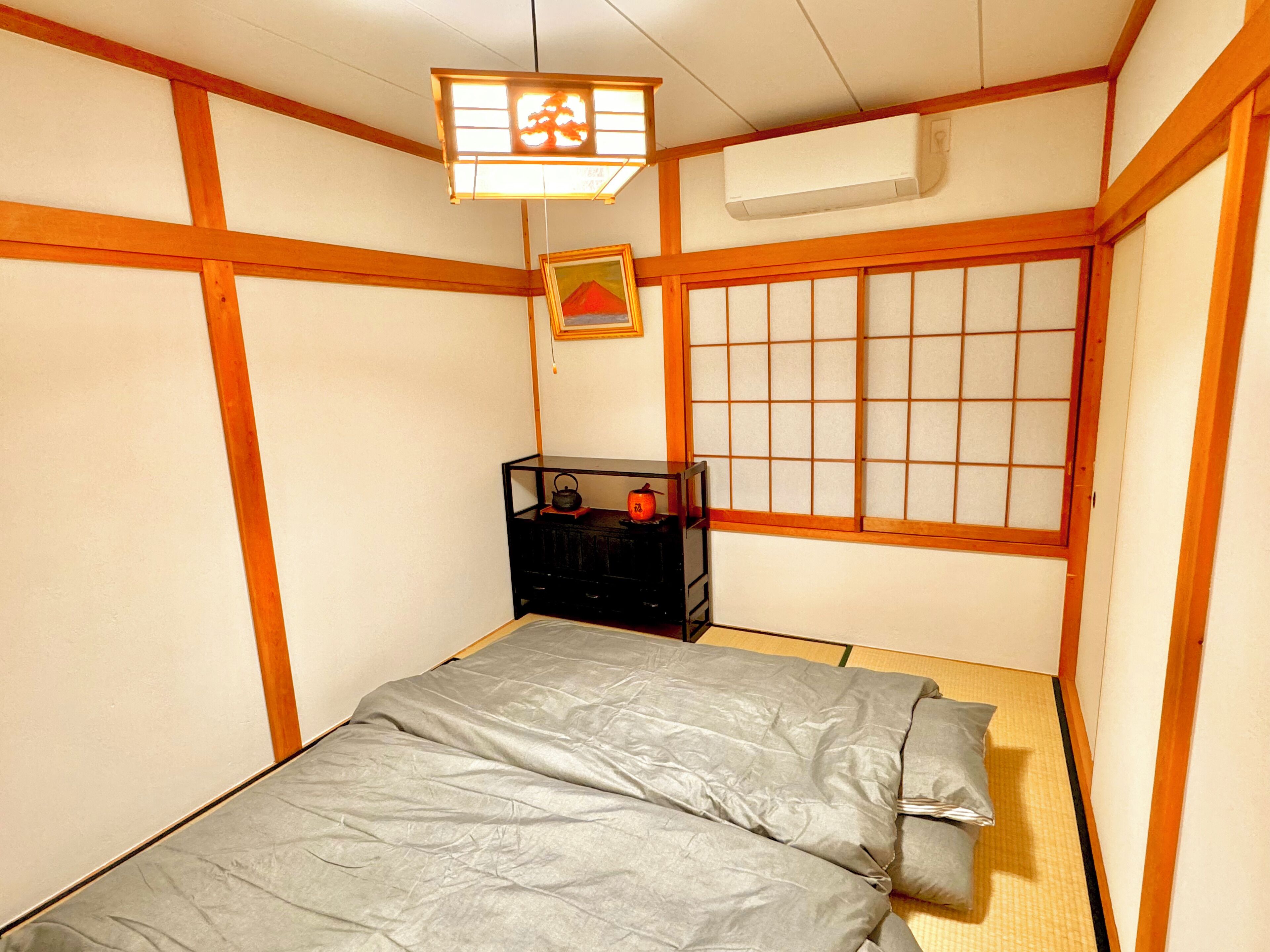 Una habitación japonesa tradicional con suelo de tatami, un futón, un shoji y un pequeño armario negro con objetos encima.