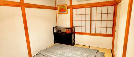 Una habitación japonesa tradicional con suelo de tatami, un futón, un shoji y un pequeño armario negro con objetos encima.
