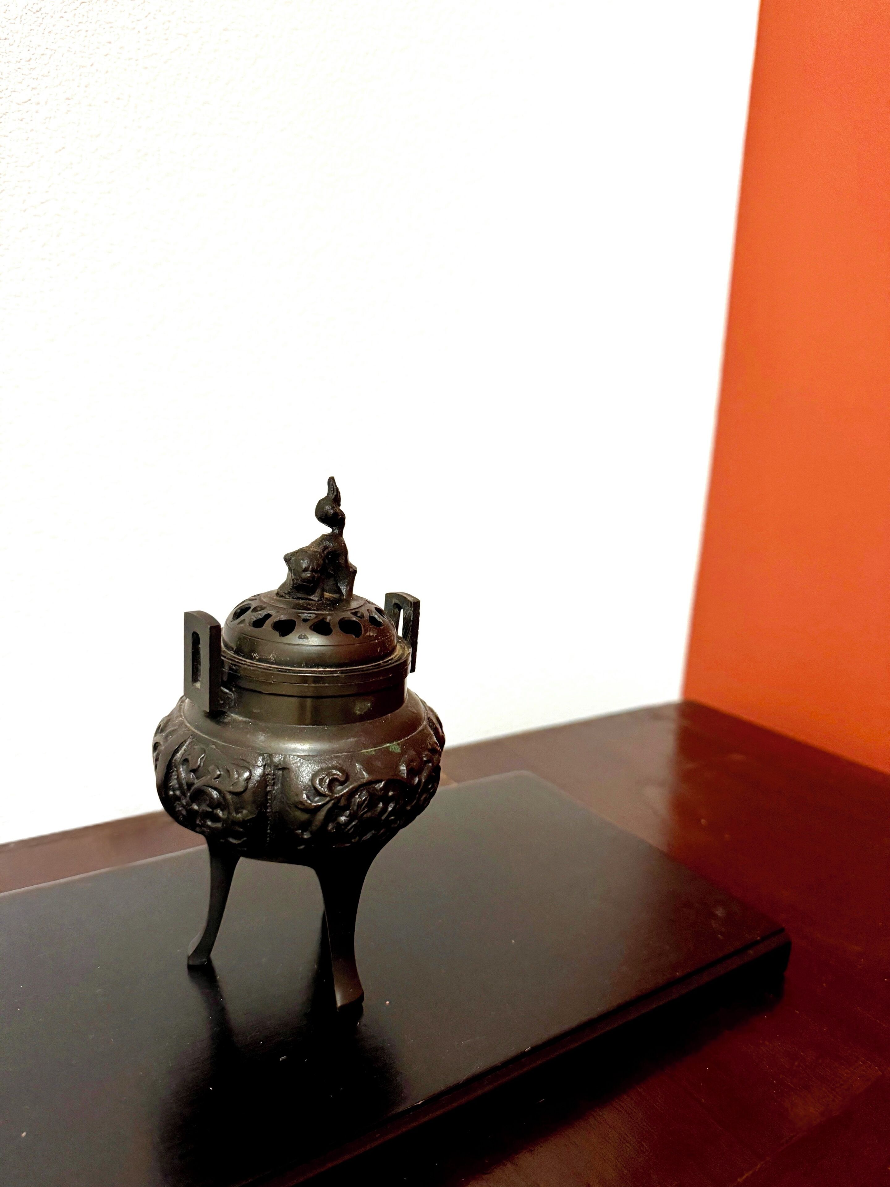 A small, ornate metal container with a lid, placed on a dark surface against a white and orange background.