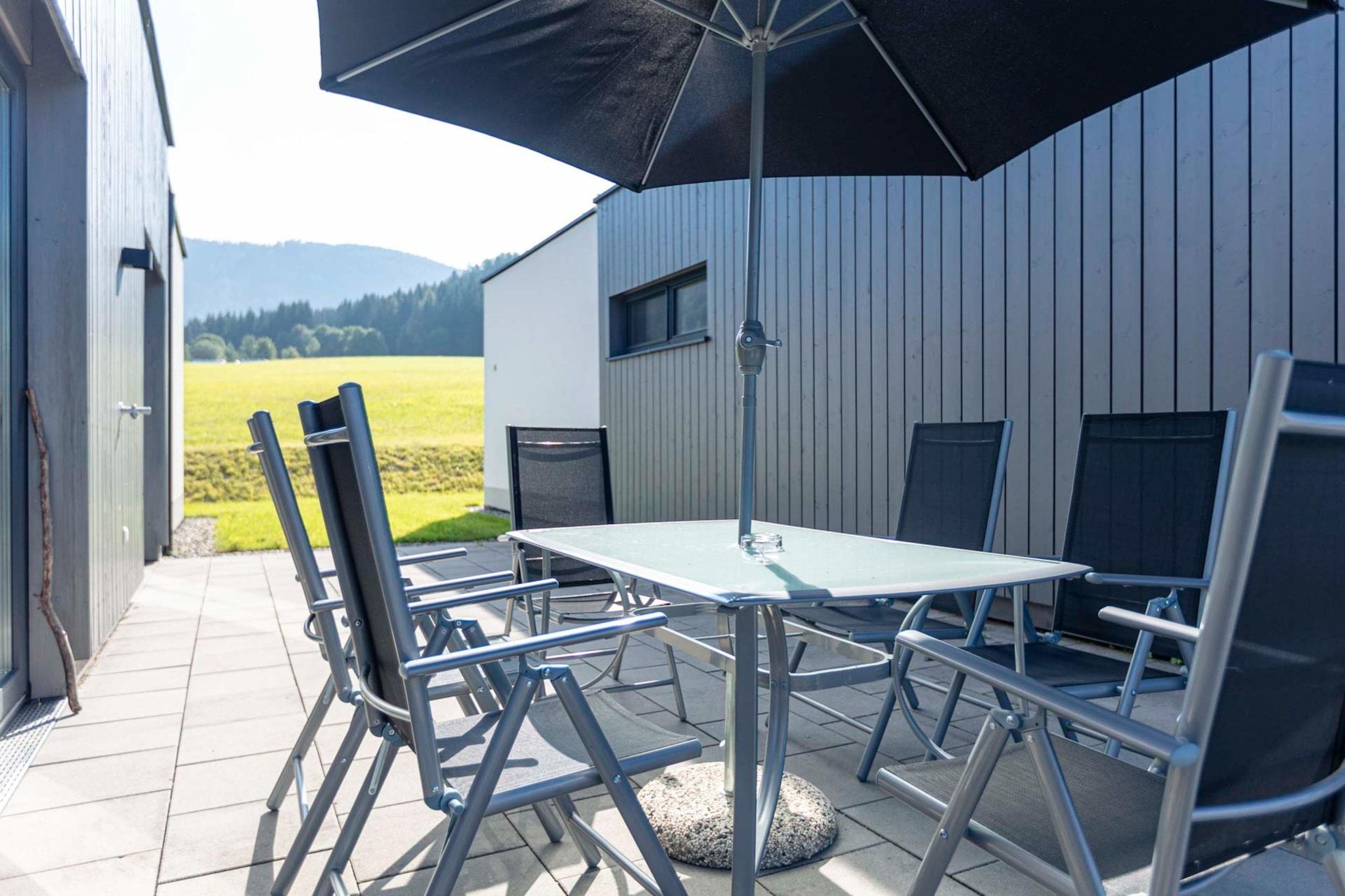 An outdoor patio with a table and chairs, a black umbrella, and a view of a field and mountains in the background.